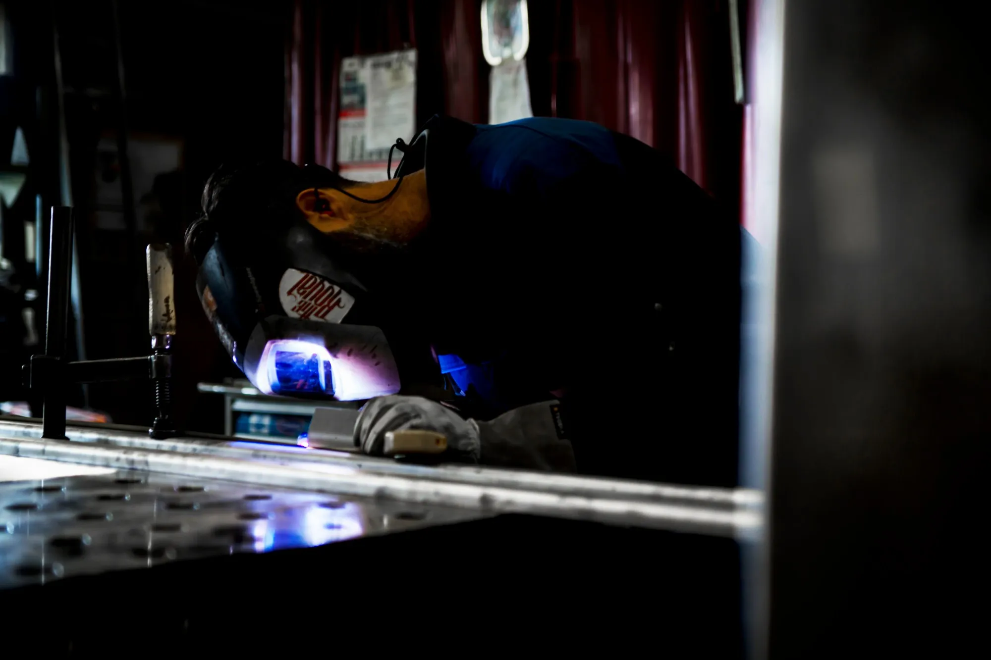 Welder in a welding helmet leaning over a long section seam clamped on the welding table